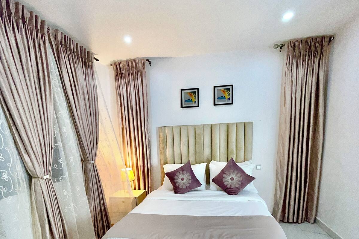 A bedroom with a bed, curtains, a headboard, and two framed pictures on the wall.