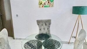 A dining area with a glass table and cushioned chairs, a green floor lamp, and a wall-mounted painting.