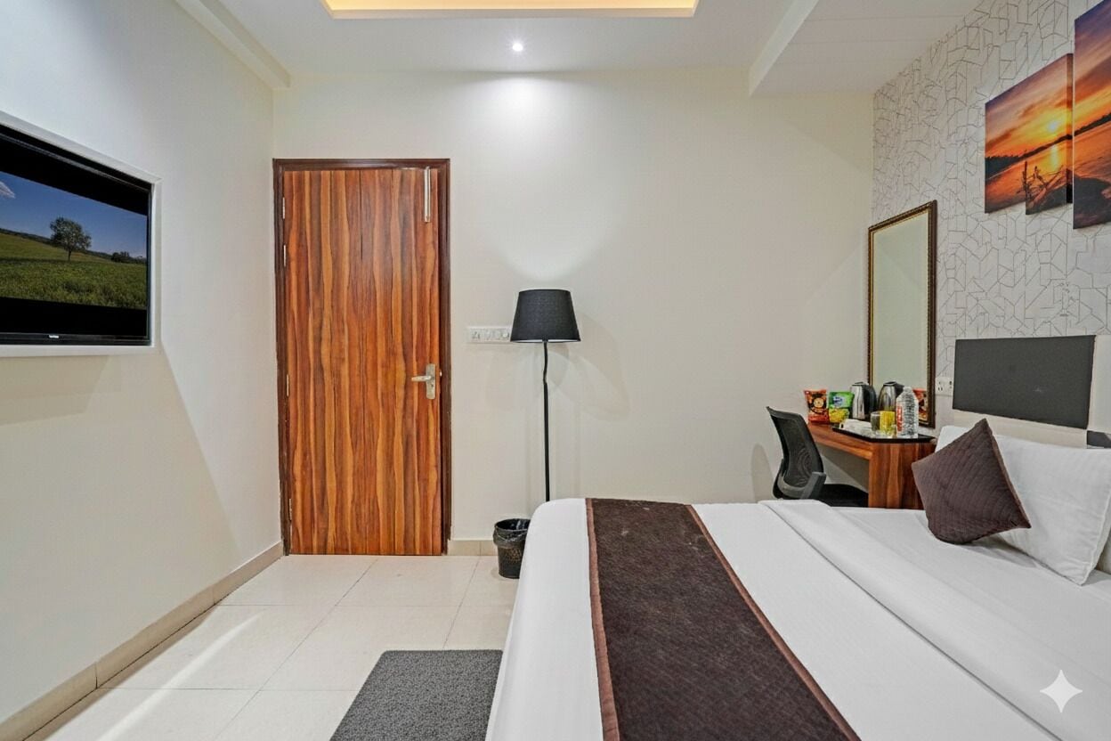 A hotel room with a wooden door, a flat-screen TV, a desk with a chair, a bed with pillows, and a wall-mounted mirror.