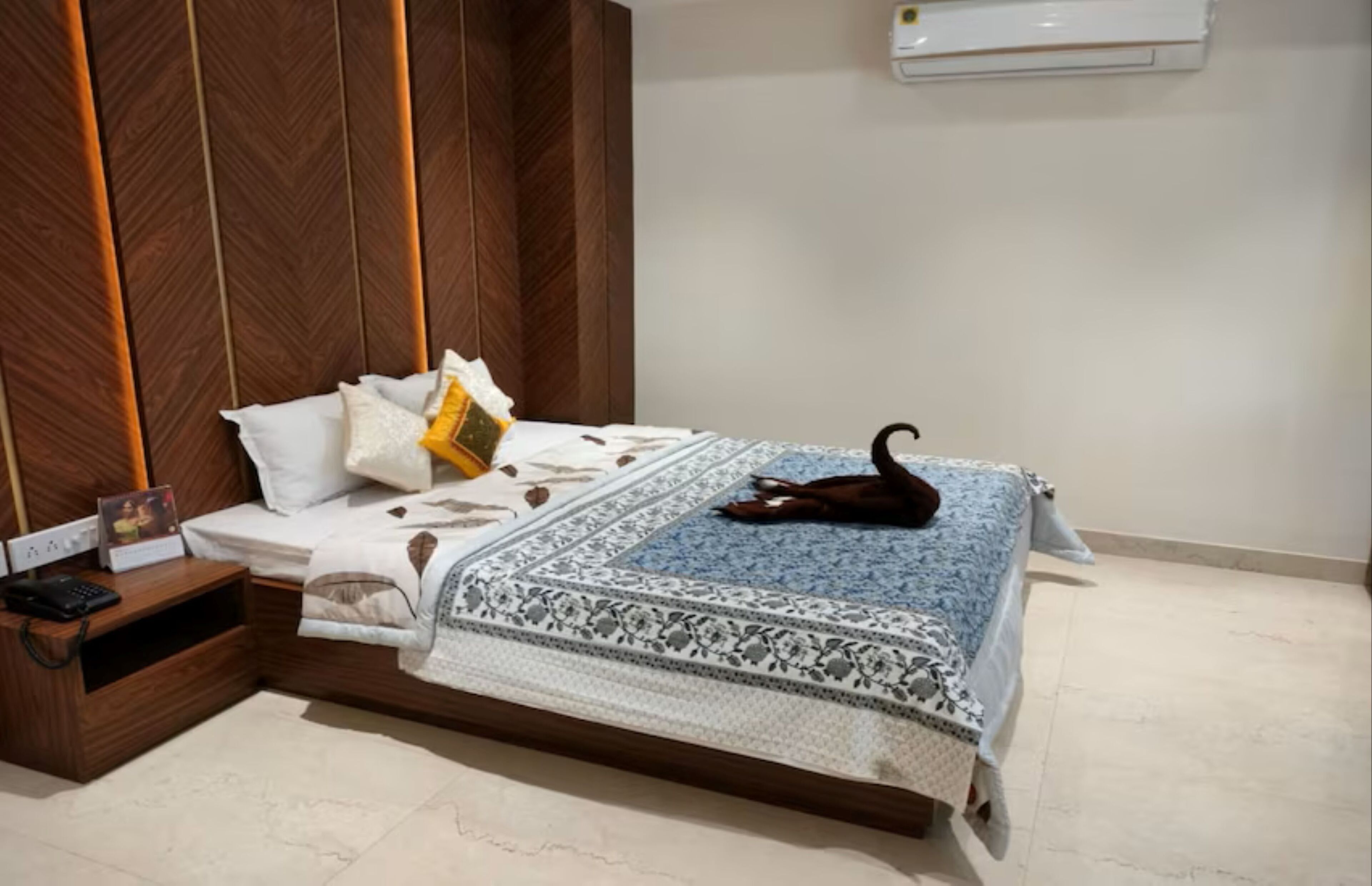 Una cama bien hecha con una colcha estampada, una mesita de noche con un teléfono y un marco de foto, y un aire acondicionado de pared.
