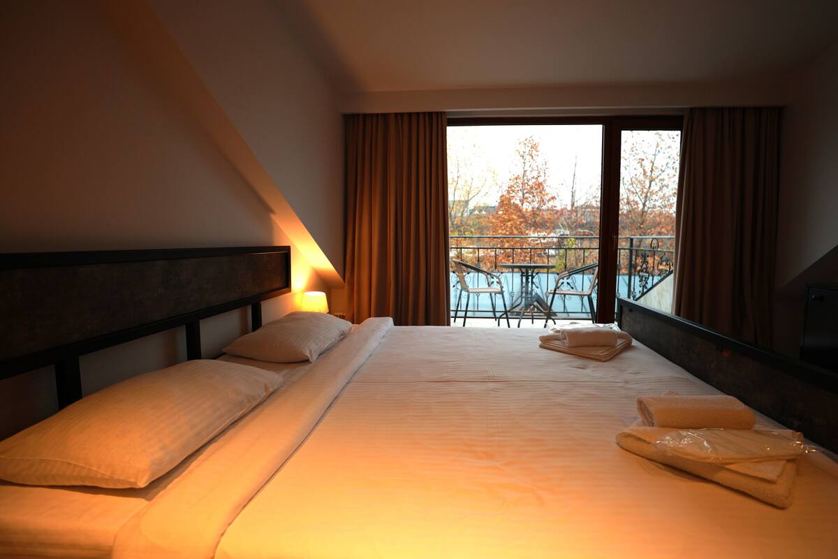 A hotel room with a large bed, a nightstand, a lamp, and a view of an outdoor balcony with a pool and chairs.