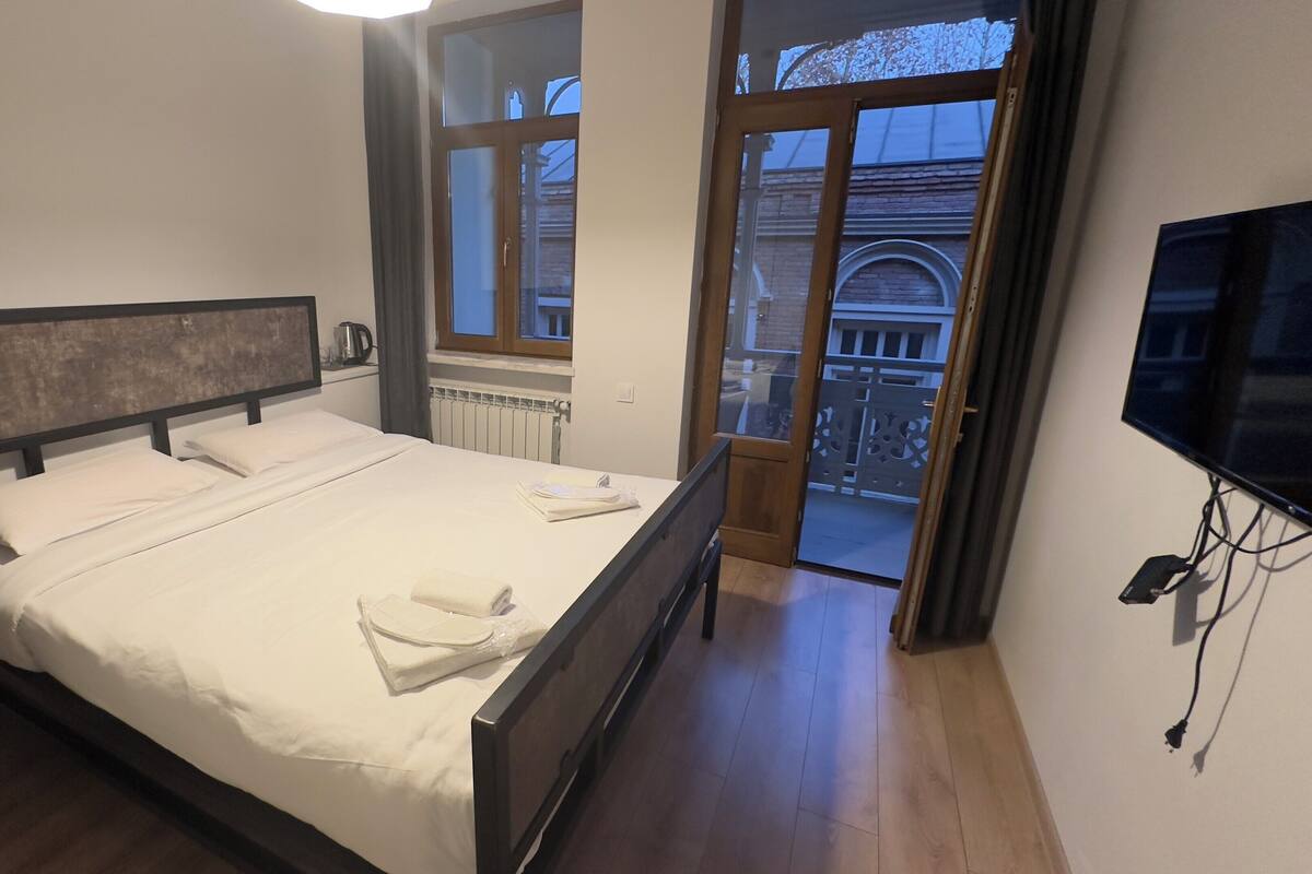 A modern hotel room with a large bed, a flat-screen TV, and a view of the outside balcony.