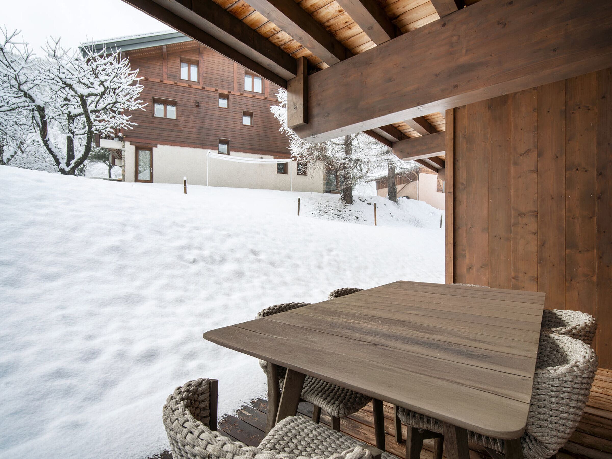 A wooden table and chairs in a snowy outdoor area with a building in the background.