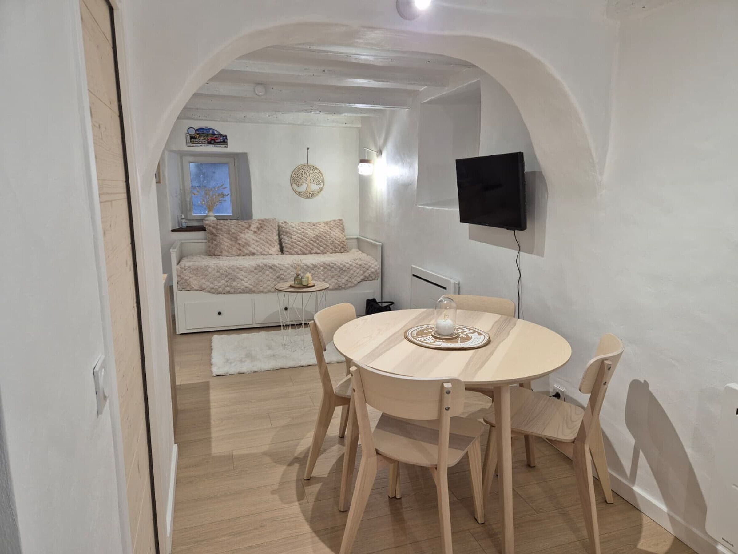 A cozy room with a round wooden table, four chairs, a TV mounted on the wall, and a bed with a window in the background.