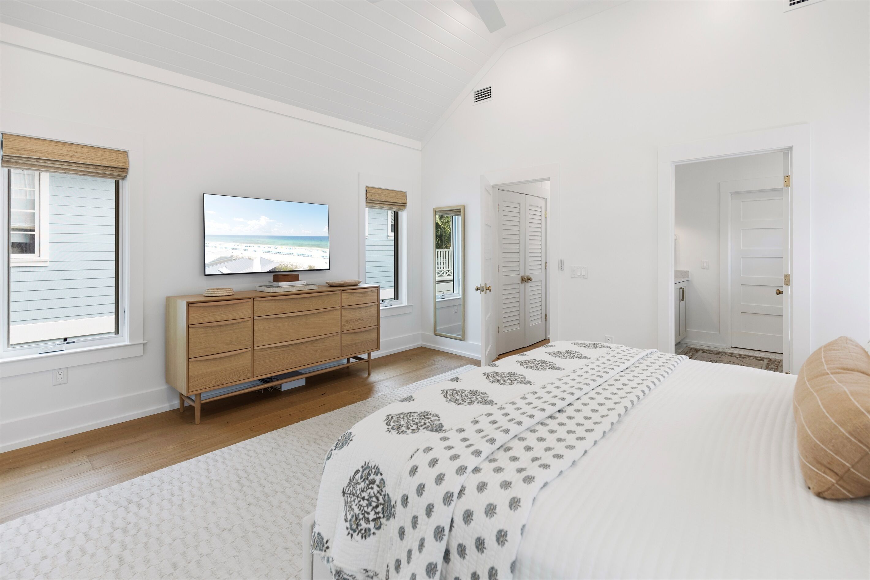 A bedroom with a large bed, a dresser, a television, and a view of the beach through the window.