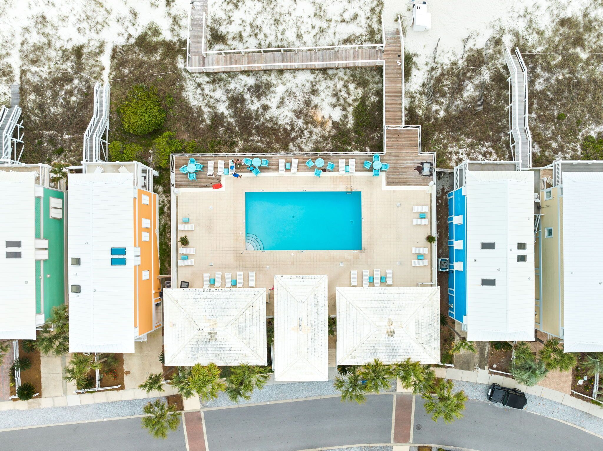 Aerial view of modern vacation property featuring central swimming pool, colorful buildings, and tropical landscaping in beach community setting.