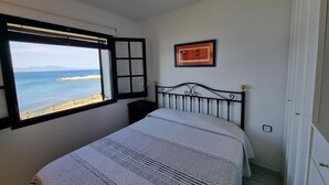 Un dormitorio con cama, ventana con vista al mar, un cuadro enmarcado en la pared y una puerta.