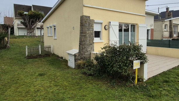 Ein Bungalow mit hellbeige Fassade, einer Steinsäule und einem kleinen Vorgarten mit einem Busch und einem Schild mit der Aufschrift 'FAVAL'.