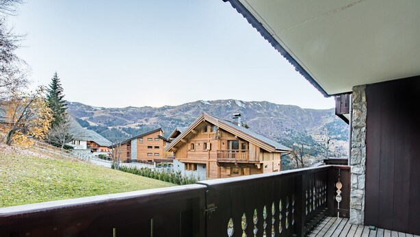 Un balcon en bois offrant une vue sur un paysage montagneux et, au loin, sur un bâtiment de style chalet.