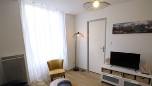 A living room with a sofa, a small table with a tray, a floor lamp, a TV on a white stand, and a door. - Cozy apartment in Luchon, 4 ppl, near spa, with parking (Bagnères-de-Luchon)