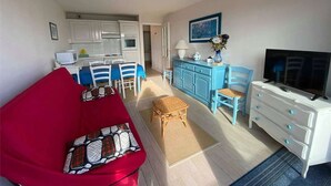 A compact living space with a red sofa, a dining area with blue chairs, a kitchen with white cabinets, a blue cabinet, a television, and a painting on the wall. - 3-Bedroom Apt near beach, with garage (Cucq-Stella Plage)
