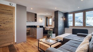 Ein modernes Interieur mit Holzboden, einer Couch, einem Glastisch, einer Küchenzeile und Blick auf eine verschneite Landschaft durch große Fenster.