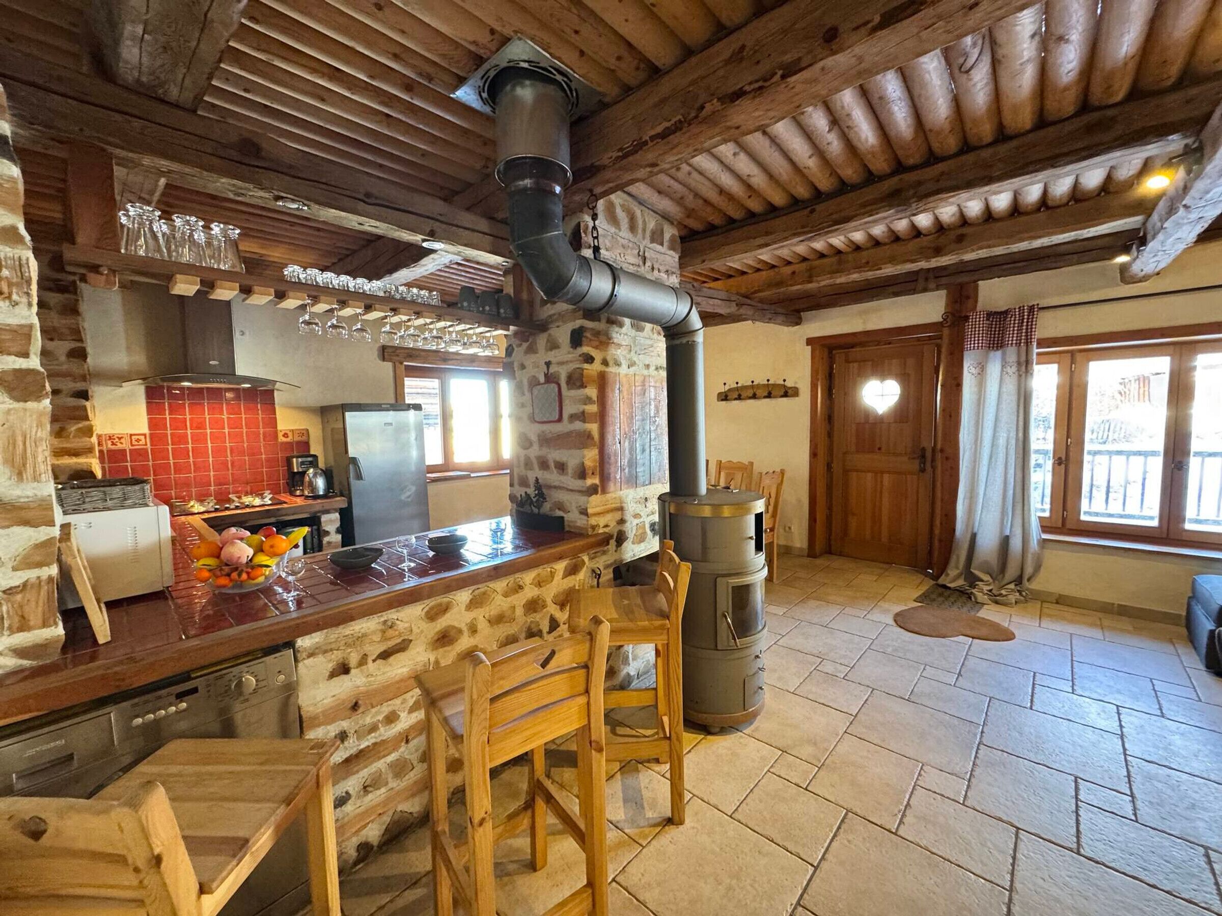 A rustic kitchen with stone walls, wooden beams, a central wood-burning stove, and a stone fireplace.