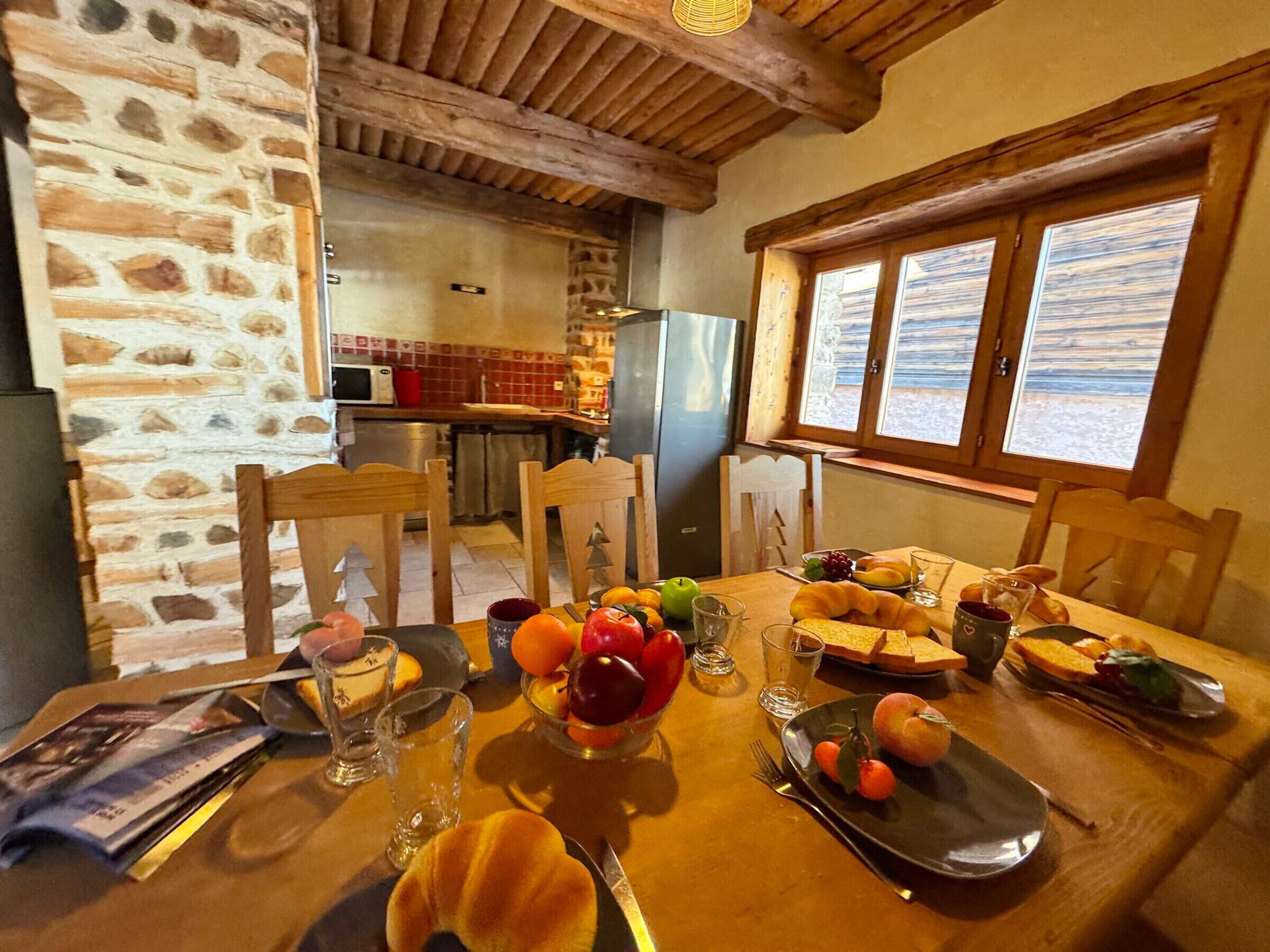 Una mesa de comedor dispuesta con frutas, pan y vasos en una habitación rústica con paredes de piedra y vigas de madera.