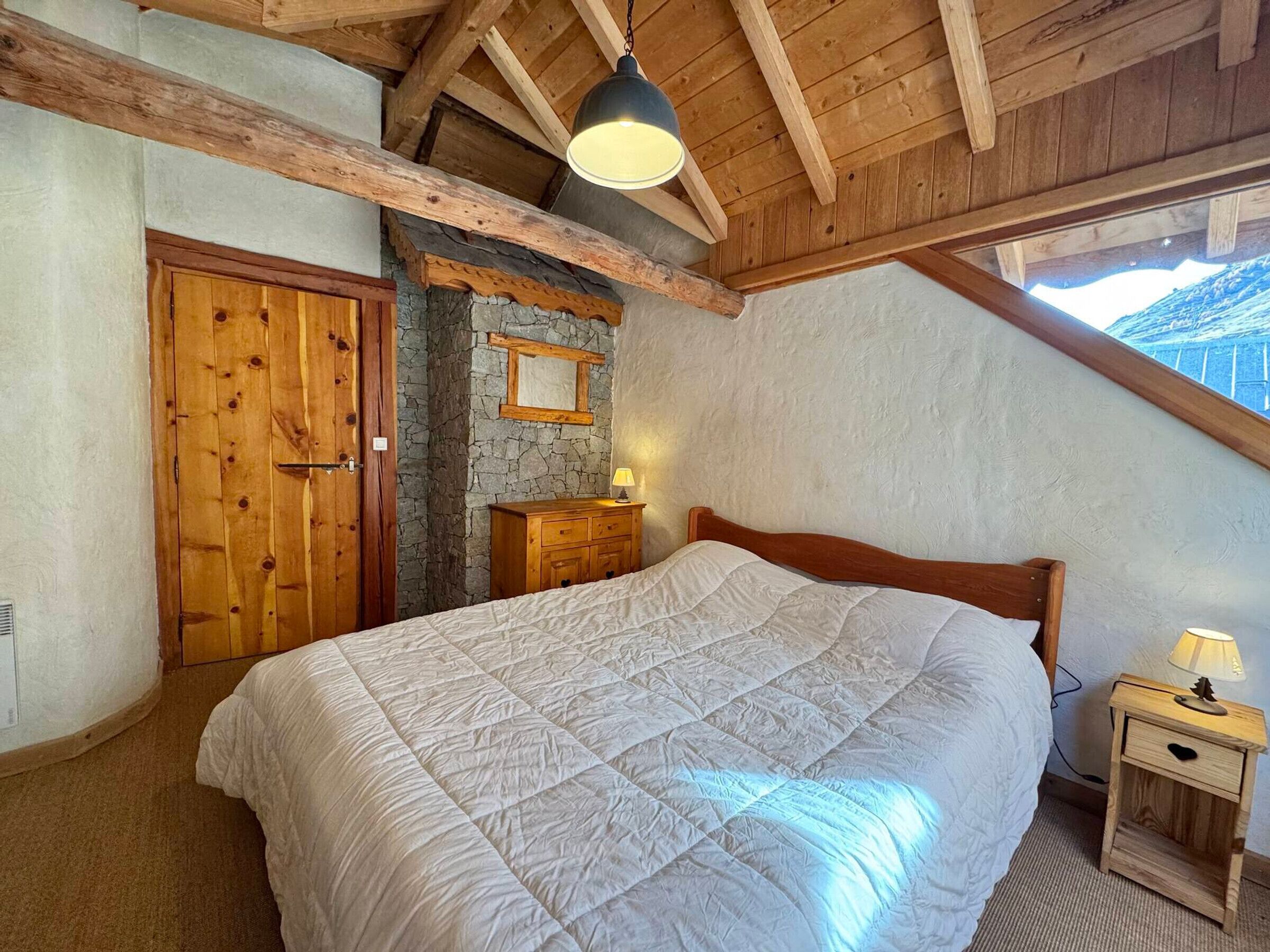 Un dormitorio con una cama grande, vigas de madera en el techo, una pared de piedra, un aparador de madera y una mesita de noche con una lámpara.