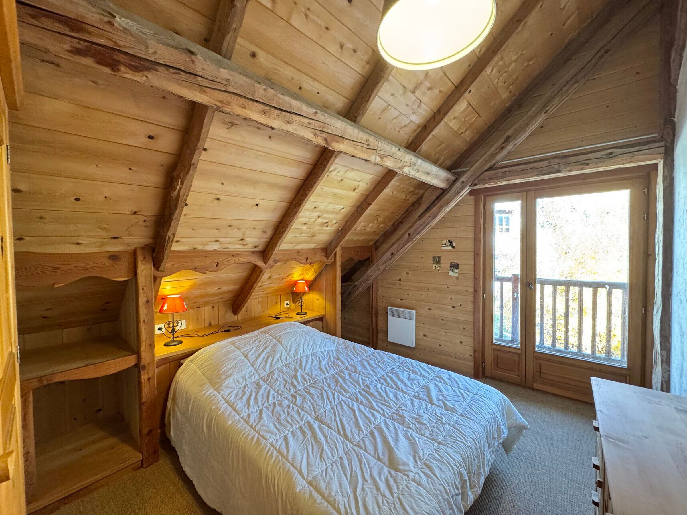 Ein Schlafzimmer einer Holzhütte mit einem Bett, zwei Nachttischlampen, einem Heizgerät und einem Fenster mit Blick auf Bäume.