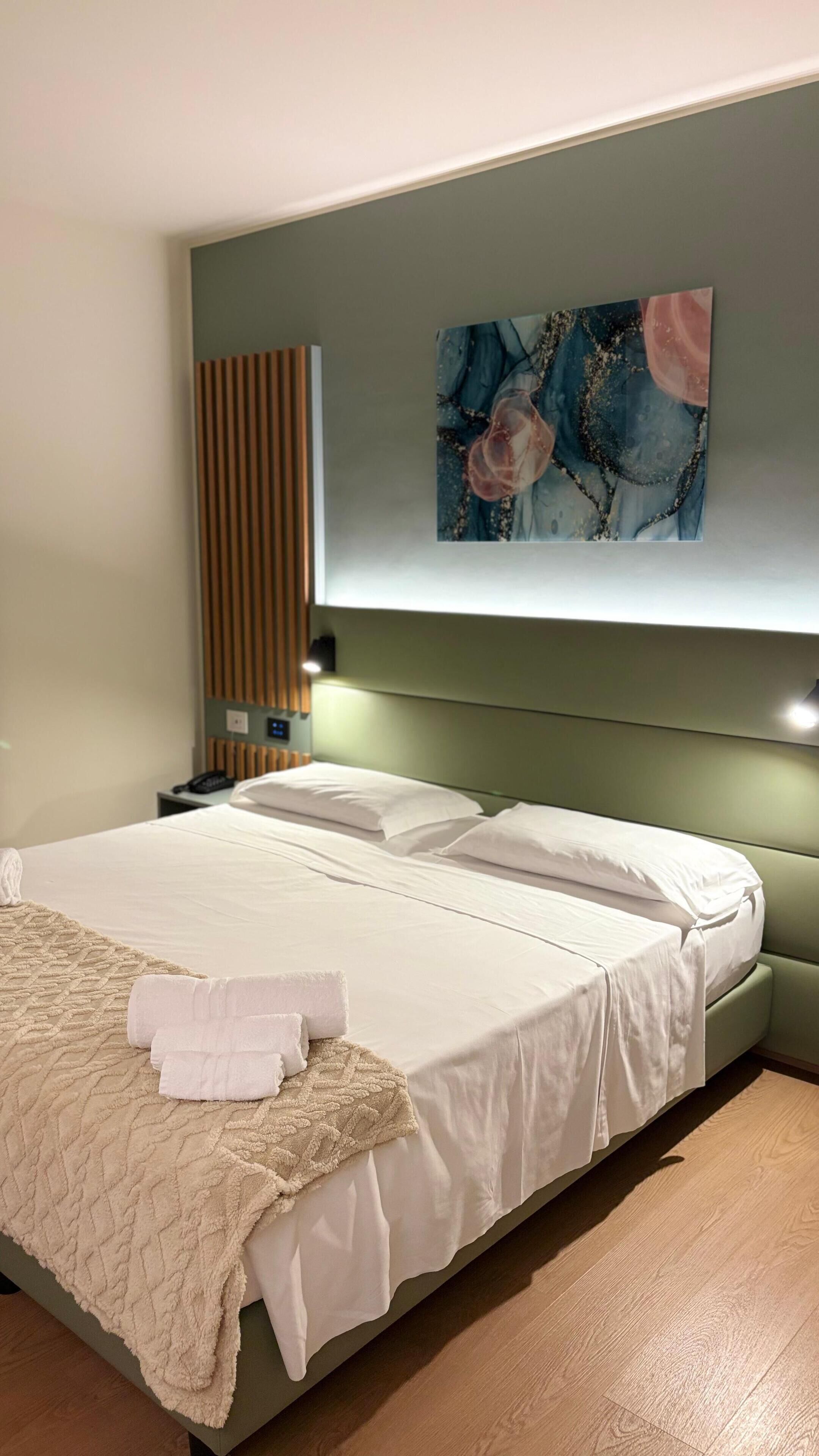 A modern hotel room with a large bed, a headboard with built-in lighting, a bedside table with a phone, and a wall-mounted artwork.