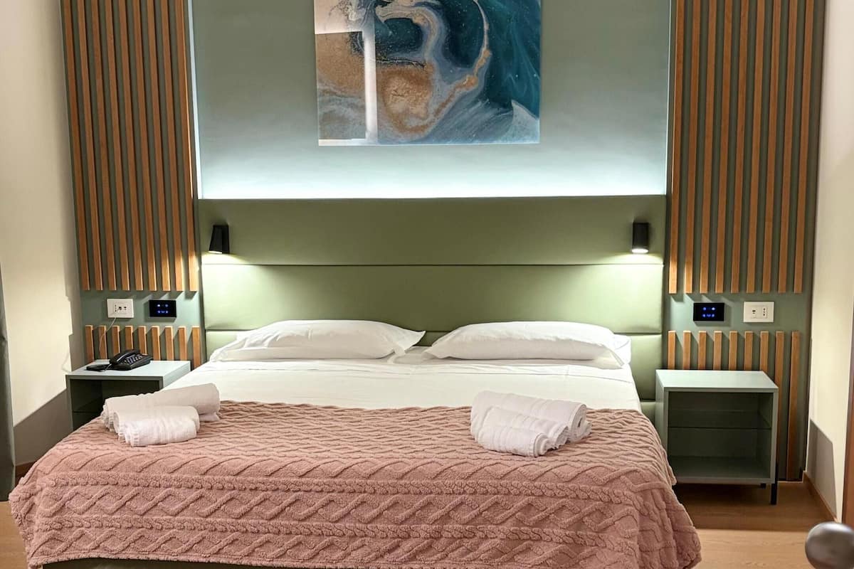 A modern hotel room with a large bed, wooden headboard, and a painting above it. There are two bedside tables with lamps and a small nightstand with a phone.