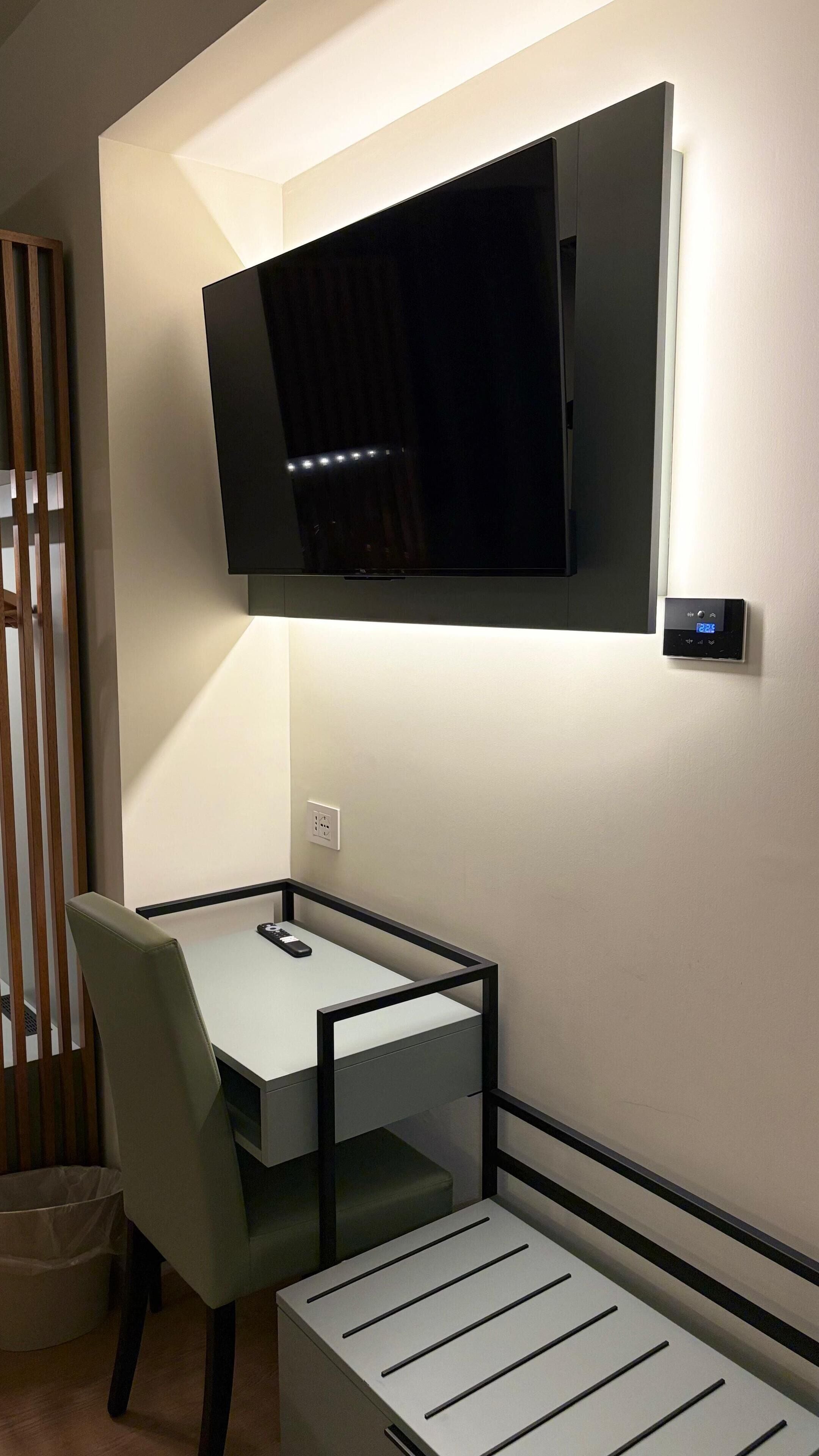 A modern room with a flat-screen TV mounted on the wall, a small table with a chair, and a trash bin.