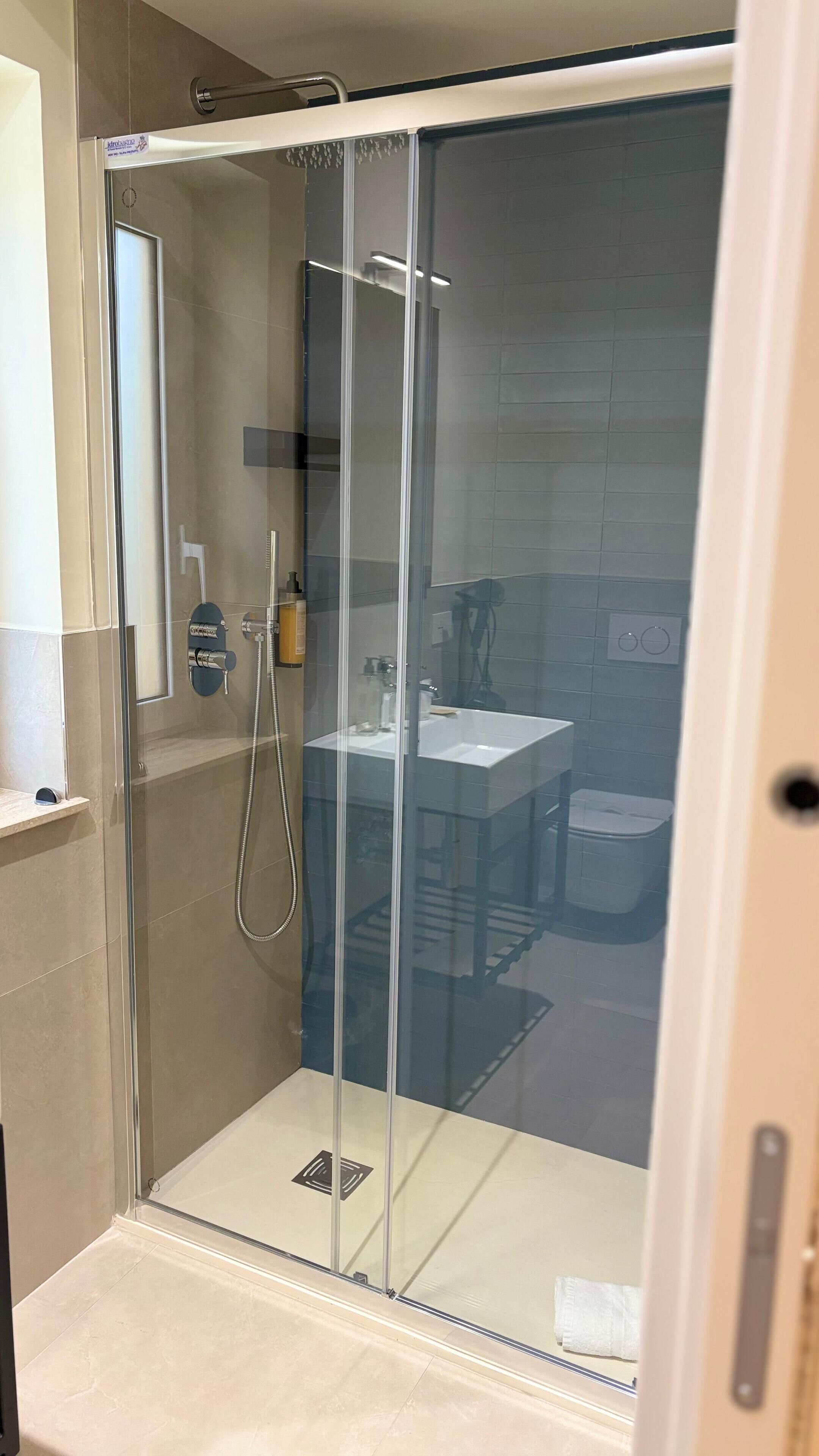 A glass-enclosed shower with a control panel, a sink, and a toilet visible through the glass.