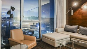 Habitación de hotel moderna con un ventanal grande que ofrece vistas a la ciudad, un sofá, una silla y una lámpara.