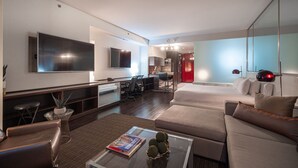 Une chambre d’hôtel moderne dotée d’une grande télévision à écran plat, d’un canapé, d’une table basse avec un livre et des objets décoratifs, ainsi que d’un coin repas agrémenté d’un mur d’accent rouge.