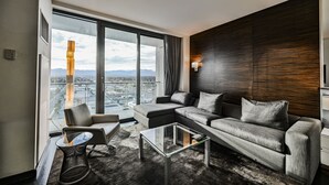 Habitación de hotel moderna con un gran ventanal que ofrece vistas a la ciudad, un conjunto de sofás, una mesa de centro de vidrio y una mesita auxiliar.