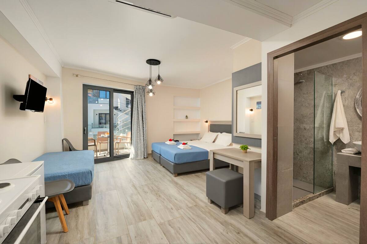 Une chambre d’hôtel moderne dotée d’une kitchenette, d’un lit recouvert d’une courtepointe bleue, d’un bureau avec une chaise, d’un petit coin salon et d’une salle de bain avec une douche en verre.