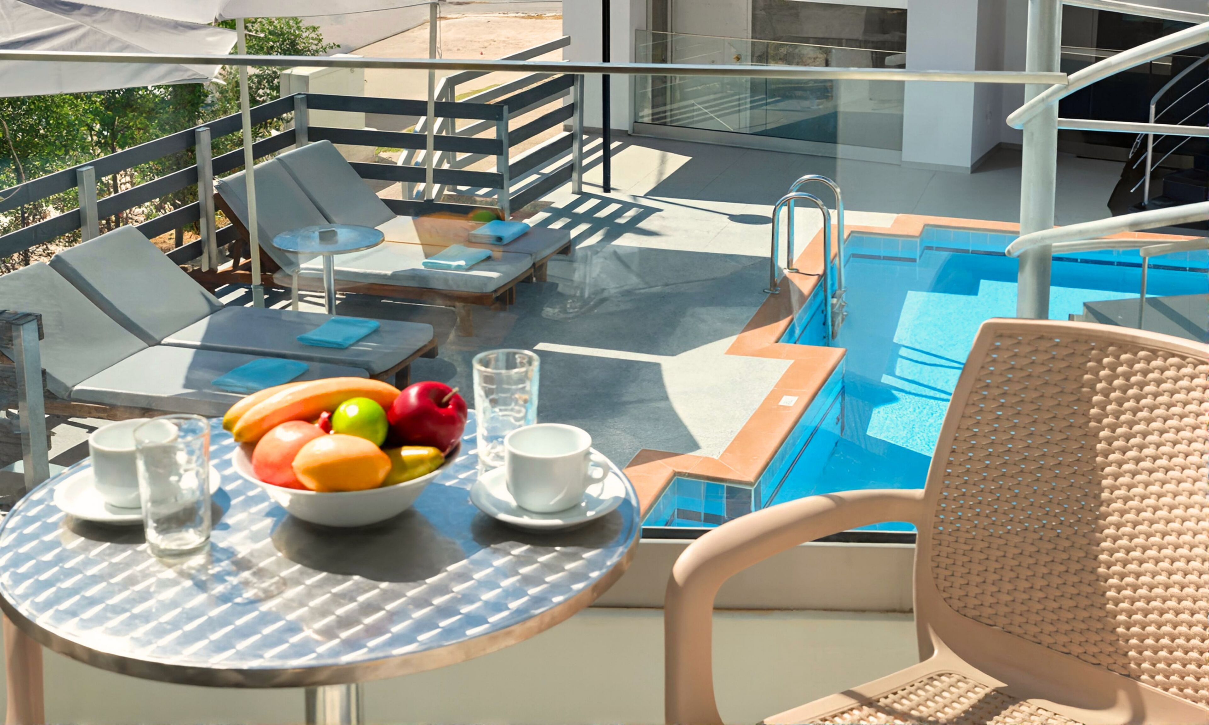 Un balcón con piscina, sillones de descanso, una mesa con frutas y bebidas, y una silla.
