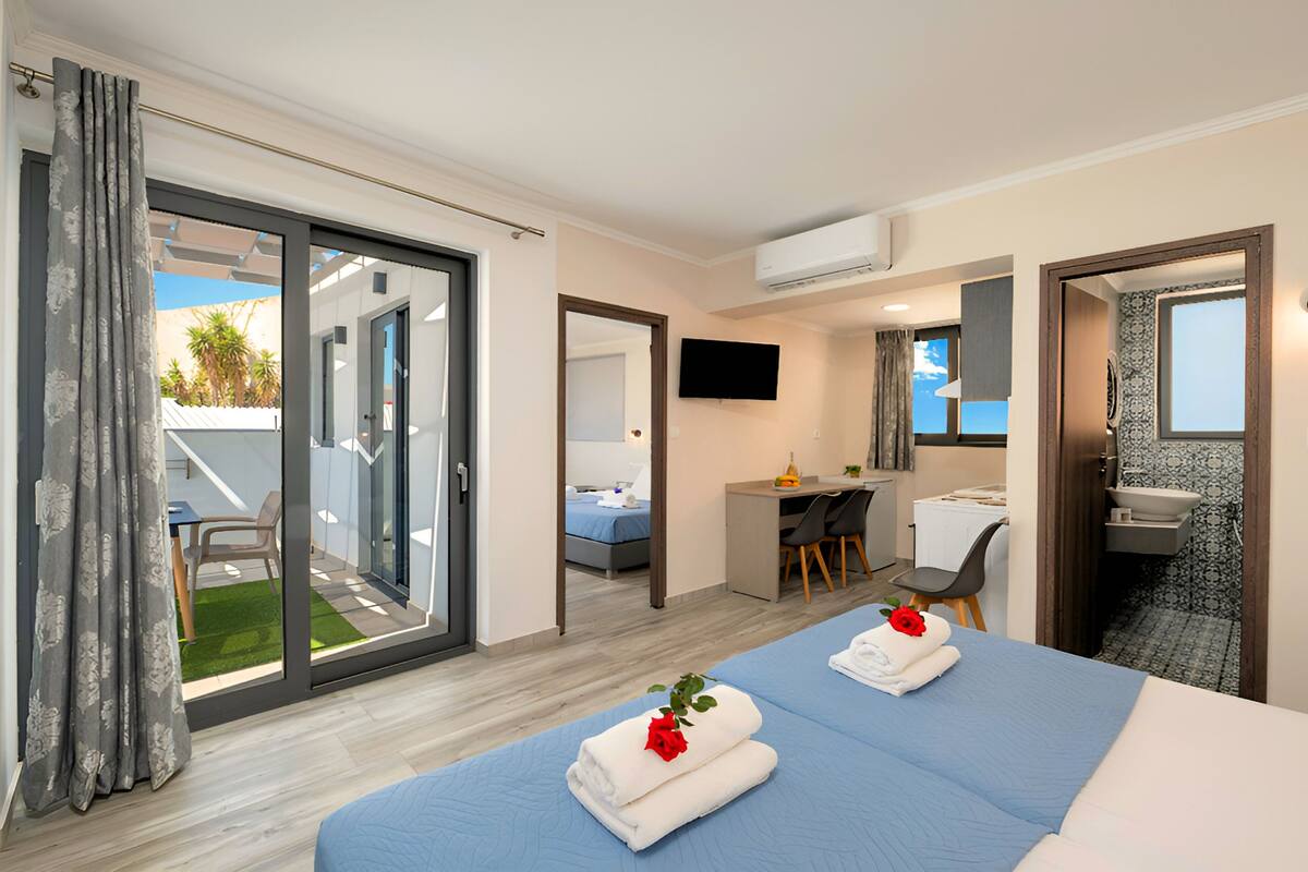 Une chambre d’hôtel dotée d’une grande porte-fenêtre coulissante donnant sur un balcon, d’un bureau avec une chaise, d’un lit avec des serviettes et une rose, et d’une salle de bain avec un lavabo et un miroir.