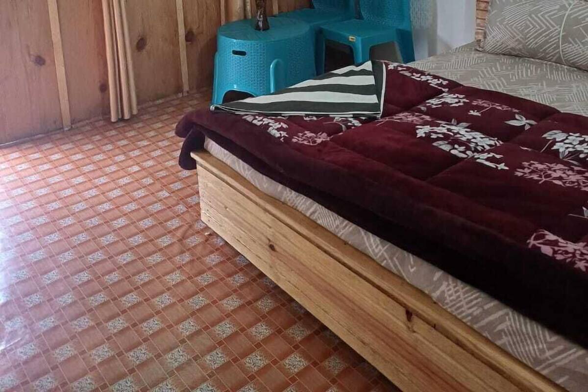 Una habitación con una cama de madera, un taburete azul y una mesita con un florero.