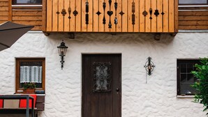 Une maison à deux étages dotée d’un balcon en bois, d’une façade blanche à texture et d’une porte marron foncé.