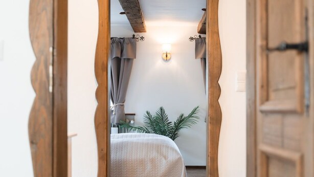 Une chambre avec un cadre de porte en bois, un lit recouvert d’une courtepointe blanche, une table de chevet et un luminaire fixé au mur.