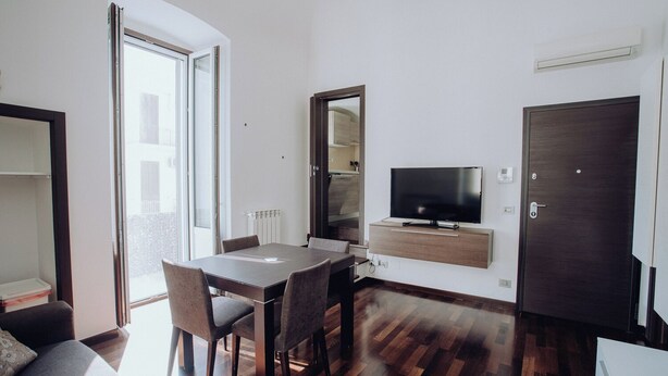Ein modernes Wohnzimmer mit Esstisch, Stühlen, einem wandmontierten Flachbildfernseher und einer Schiebetür, die auf einen Balkon führt.
