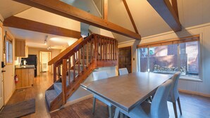 A spacious dining area with a wooden staircase leading to an upper level, a kitchen in the background, and a view of a stone wall through large windows. - The Heavenly Foothills For 4 (South Lake Tahoe)