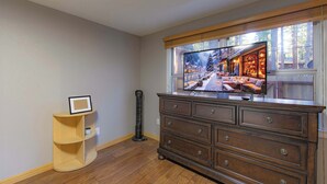 A room with a large window displaying a snowy village scene, a wooden cabinet with drawers, a small wooden shelf, and a tall black object. - The Heavenly Foothills For 4 (South Lake Tahoe)