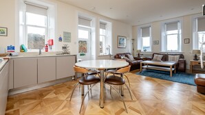 Ein geräumiger Wohnbereich mit einem runden Esstisch, einer Couch und einem Couchtisch. Große Fenster sorgen für natürliches Licht und einen Ausblick ins Freie.