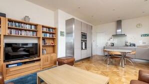 Ein modernes Wohnzimmer mit Flachbildfernseher, Holzbücherregalen, einem Couchtisch und einem Essbereich mit rundem Tisch und Stühlen.