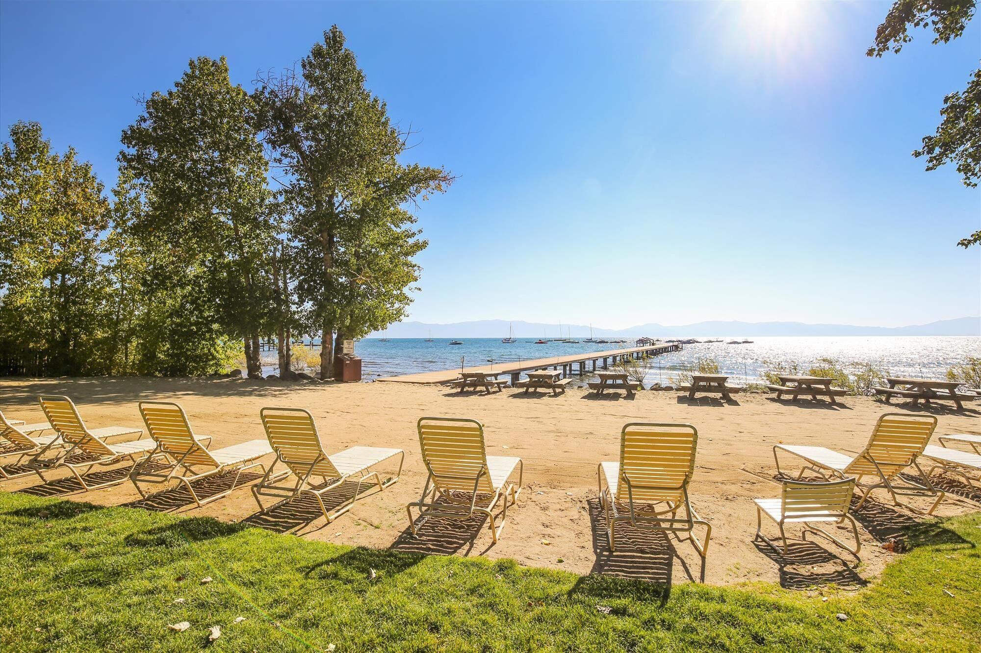A beach with rows of sun loungers and picnic tables, a wooden pier extending into the water, and a clear sky.
