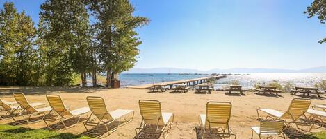 A beach with rows of sun loungers and picnic tables, a wooden pier extending into the water, and a clear sky.