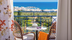 A balcony with wicker chairs, a small table, and a view of a coastal town and sea. - Astra Village (Koutouloufari)