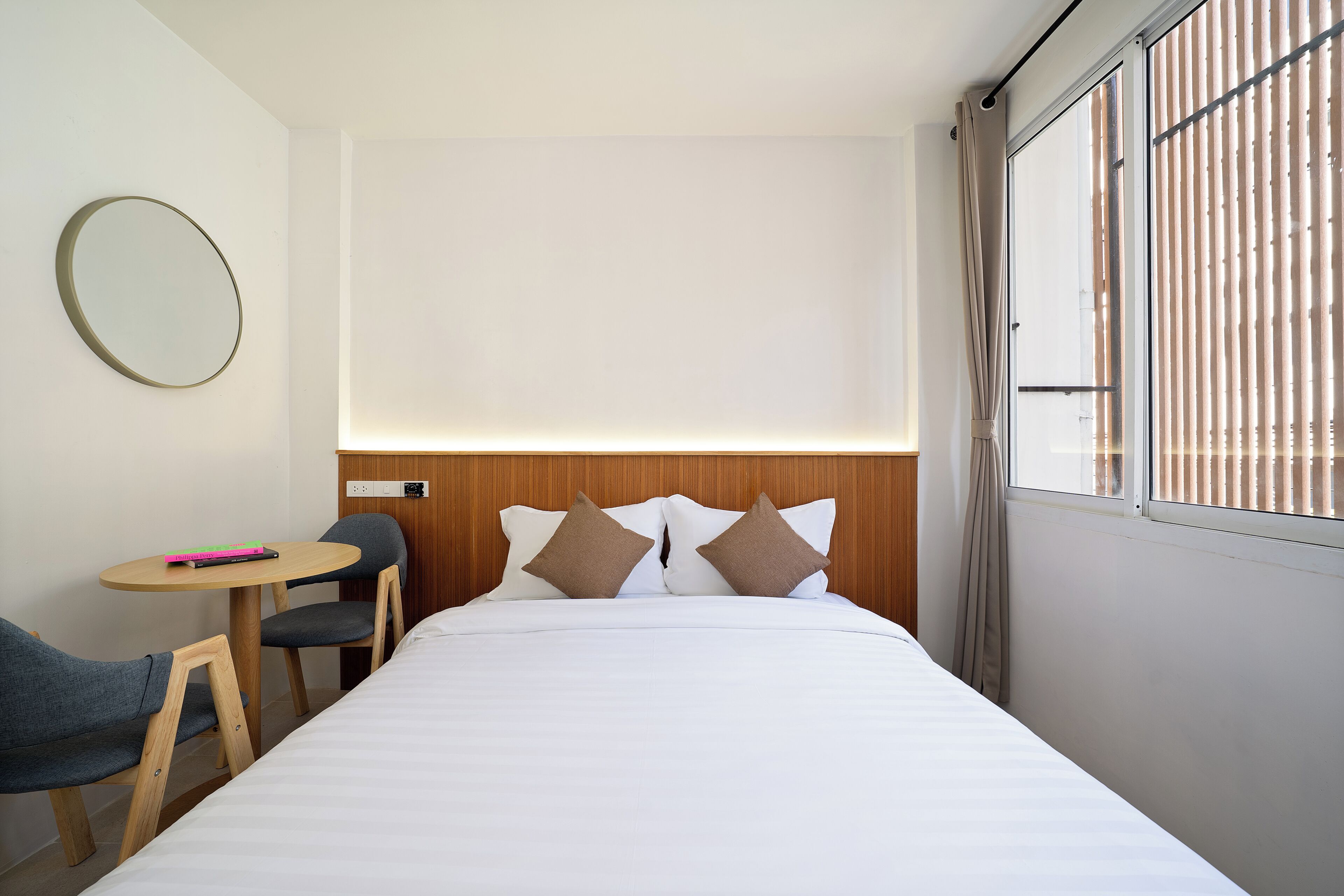 Una habitación de hotel con cama, un espejo redondo, una mesita con una silla y una ventana con persianas.