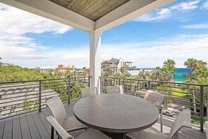 Un área de estar techada al aire libre con una mesa y sillas, con vista a una playa con palmeras y un cielo despejado.