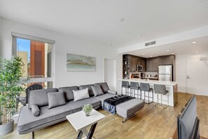 A modern living room with a grey sofa, a small table, and a TV. It includes a kitchen area with a fridge, stove, and bar stools. - Stylish 2BR Deluxe Family Apartment by Dtla (Los Angeles)