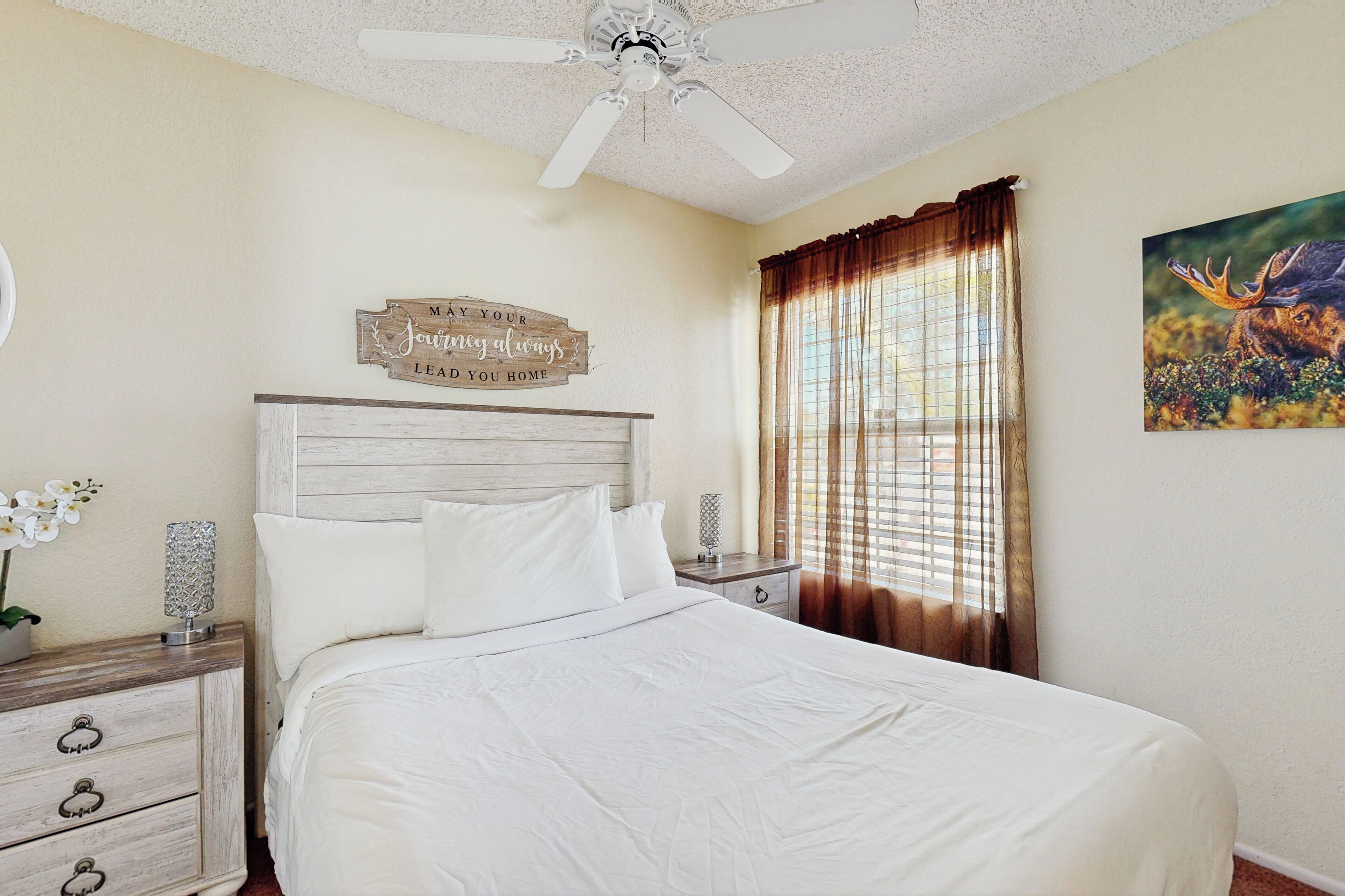 A bedroom with a bed, wooden headboard, a nightstand with a lamp, a decorative sign, and a window with curtains.