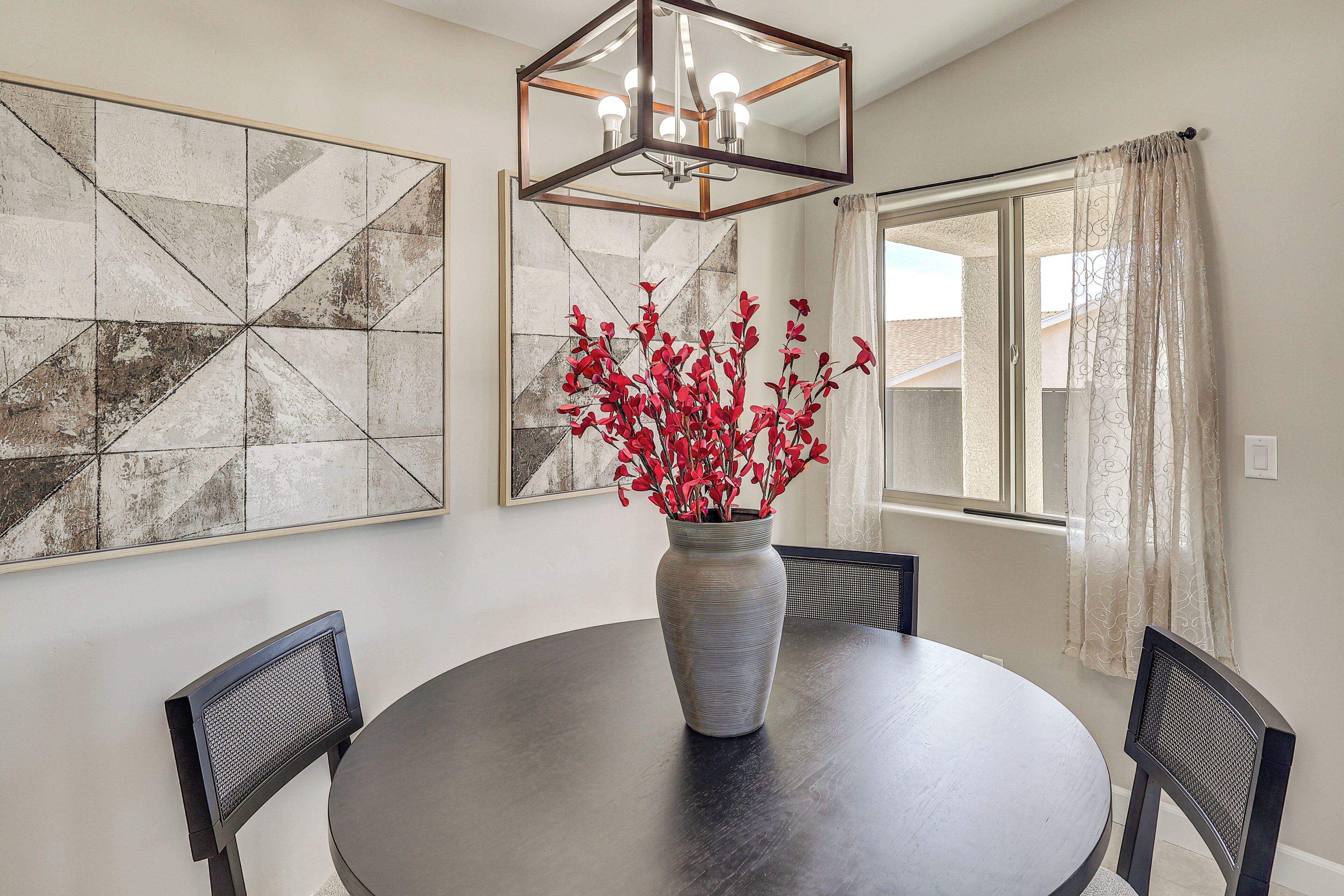 A dining area with a round table, two chairs, a vase with red flowers, a geometric wall art piece, and a window with curtains.