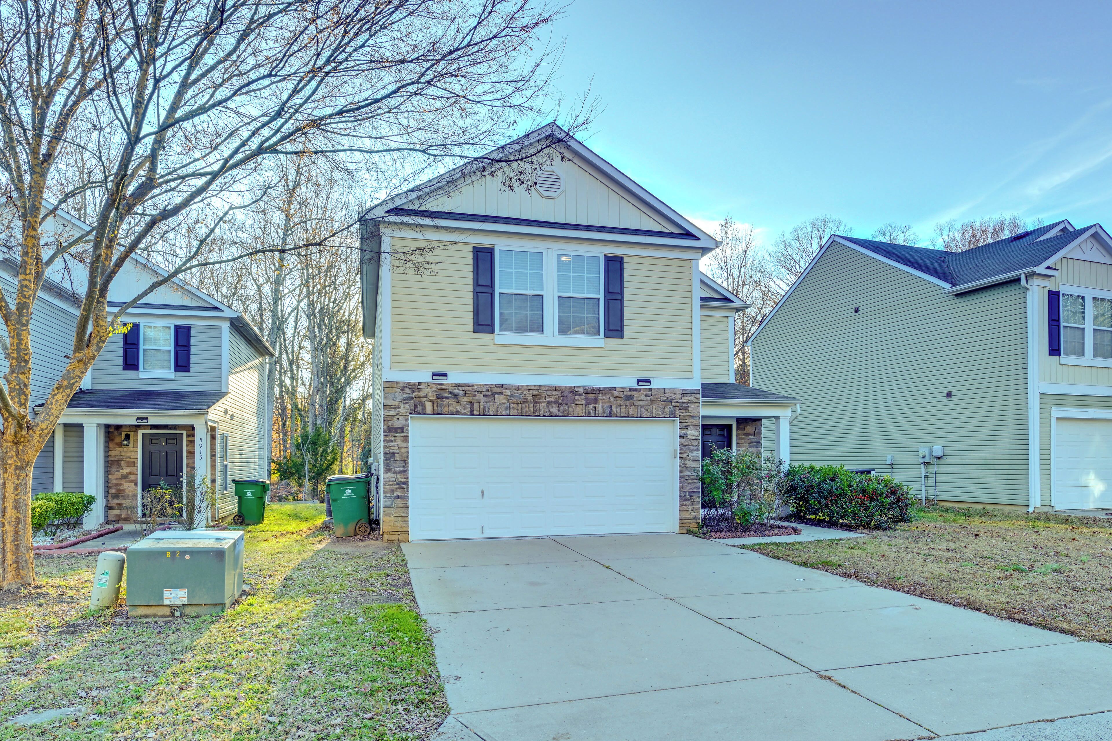 A two-story house with a garage, a stone accent wall, and a front yard with a tree and two green trash bins.