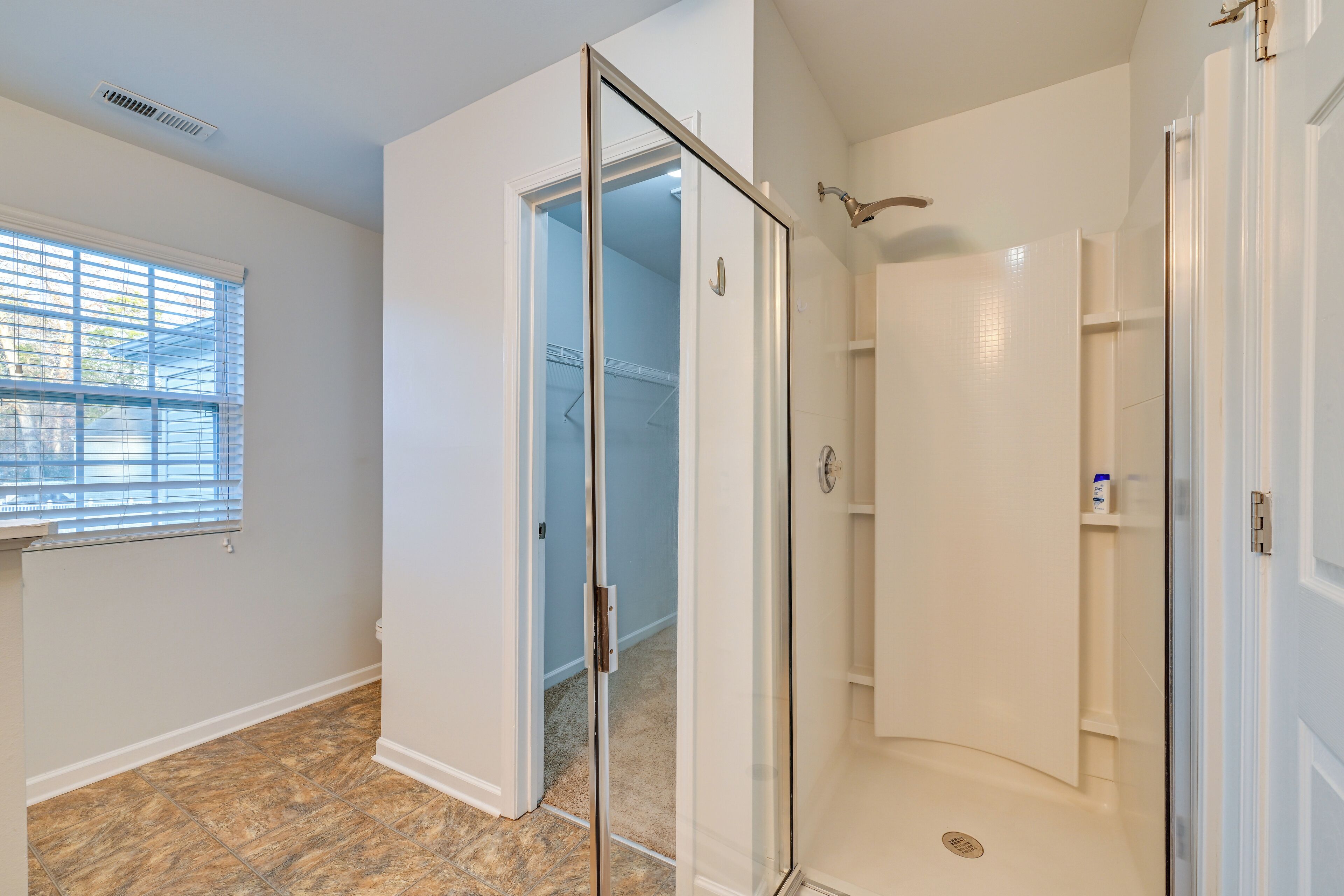 Un baño con cabina de ducha de vidrio, vestidor y ventana con persianas.