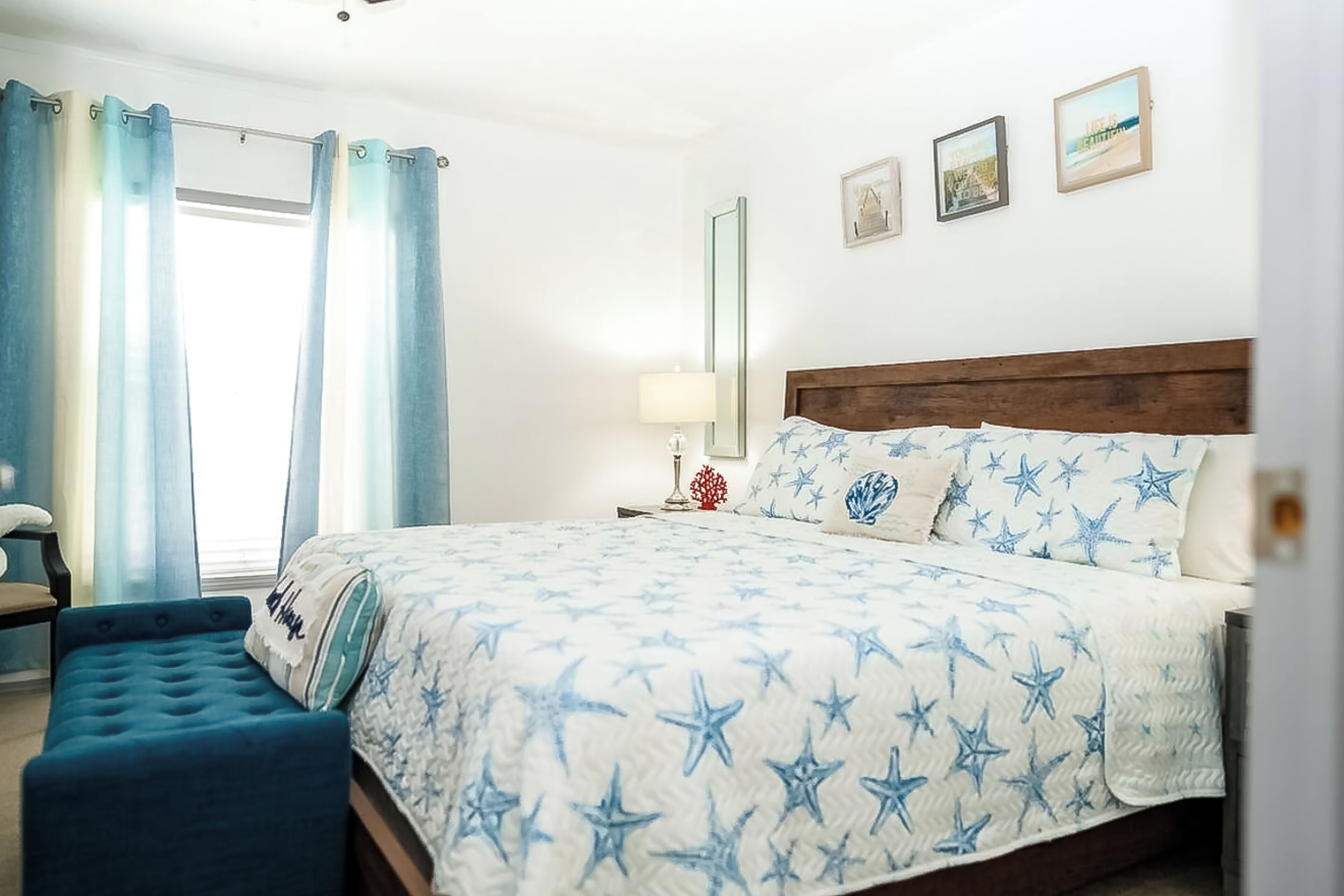 A bedroom with a bed, a bench, a nightstand, a lamp, and a window with curtains.