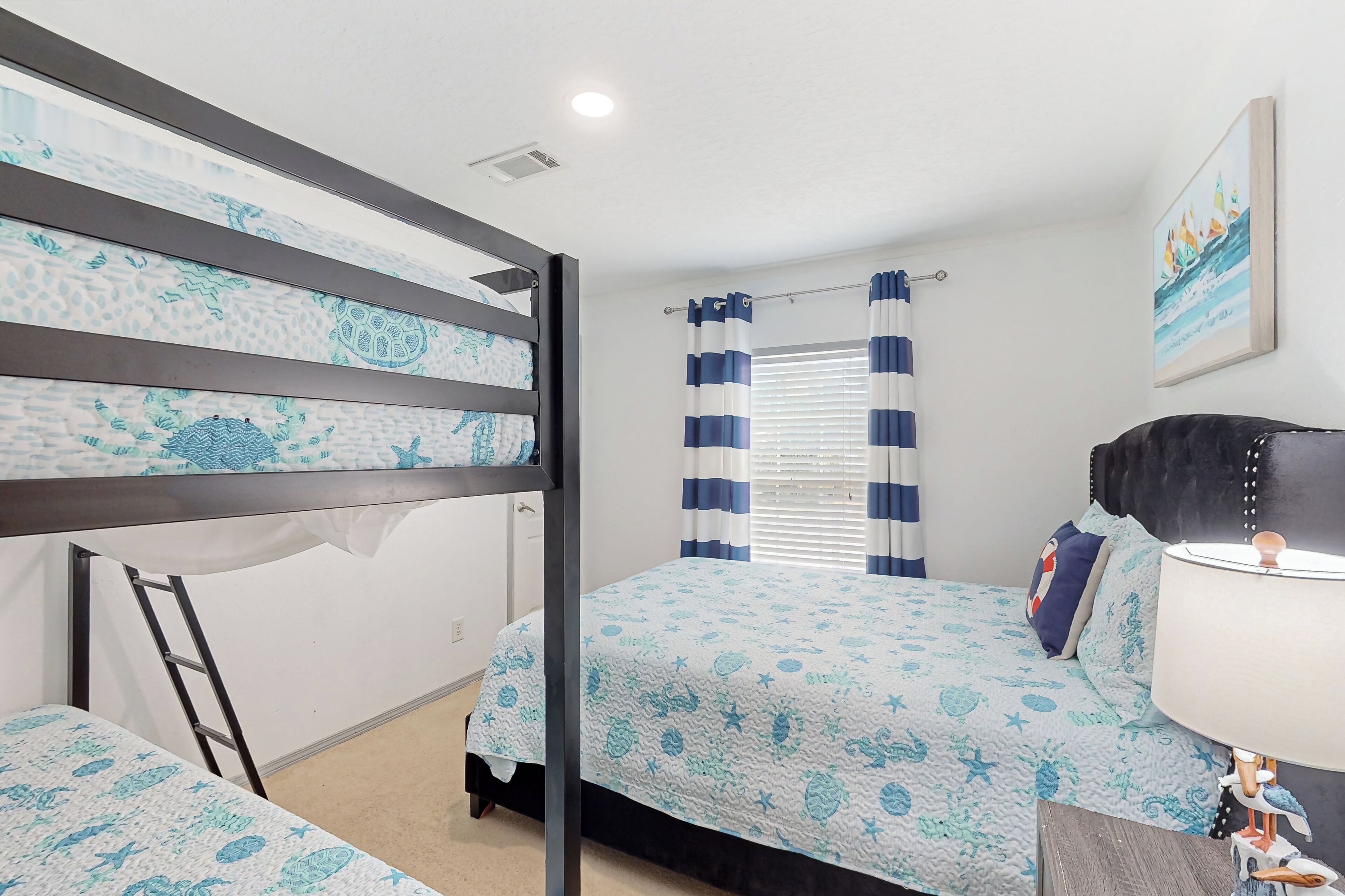 A bedroom with a bunk bed, a single bed, a window with curtains, a bedside lamp, and a wall-mounted picture.